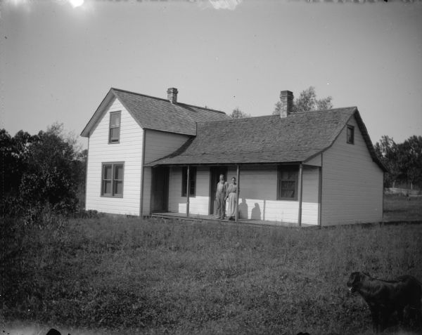 Outdoor view across lawn of a man and woman posing standing on the porch of a wooden house. There is a dog standing in the foreground on the right.