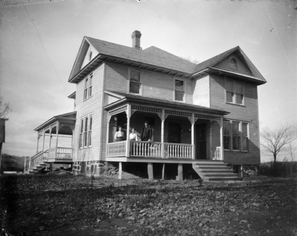 Outdoor view across lawn towards a man and two women posing standing on the porch of a two-story wooden house.