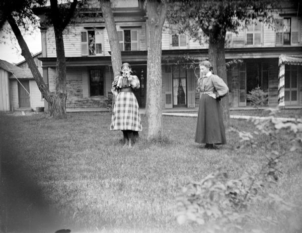 Outdoor view of two European American women posing standing in the yard under trees in front of a large two-story house.