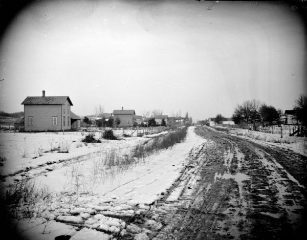 View down unpaved street towards several buildings and snow-covered fields.