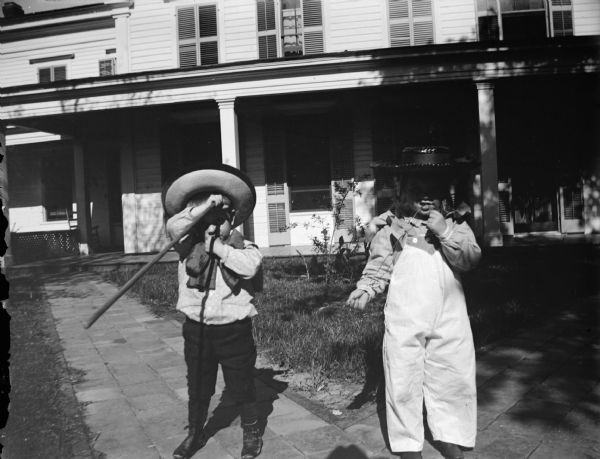 Outdoor portrait of two European American boys wearing hats. The boy on the left is holding a stick, and the boy and the right is holding a string. They are posing standing on a brick walkway in front of a two-story wooden house.