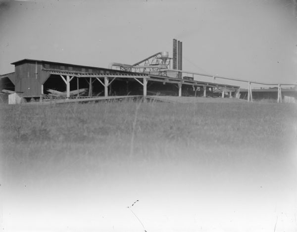 View across field towards a long industrial building with four smokestacks. Probably a lumber mill with a saw dust burner.