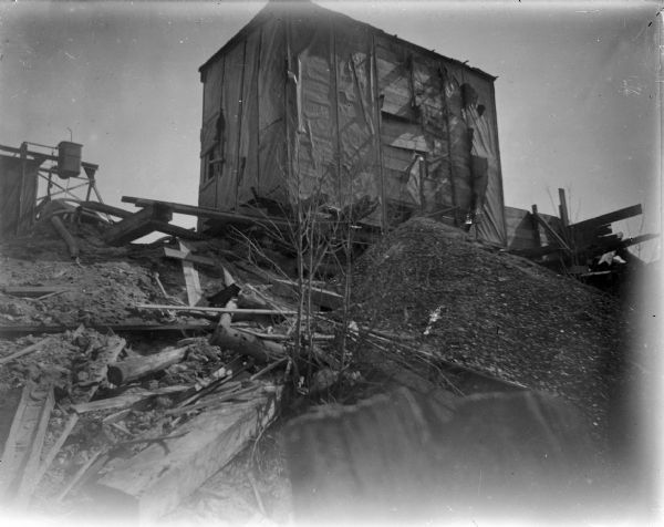 View looking up towards a small building standing at the top of a slope. Equipment and piles of building material surround the tar paper shack. Probably a tar paper shack at a construction site during the rebuilding after the flood of 1911.