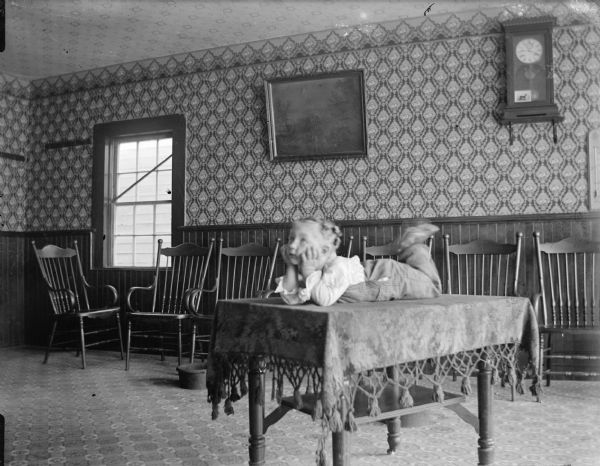 Indoor portrait of a European American boy posing lying on his stomach on top of a cloth covered table with his head in his hands, possibly taken inside the Merchant's Hotel.