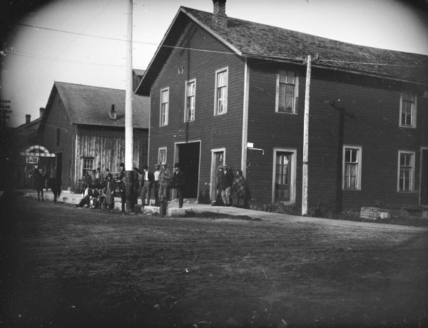 View across unpaved street towards a group of men and women, probably Native Americans, posing sitting and standing in front of Building #88 on the northeast corner of First and Fillmore Streets and the City Livery.