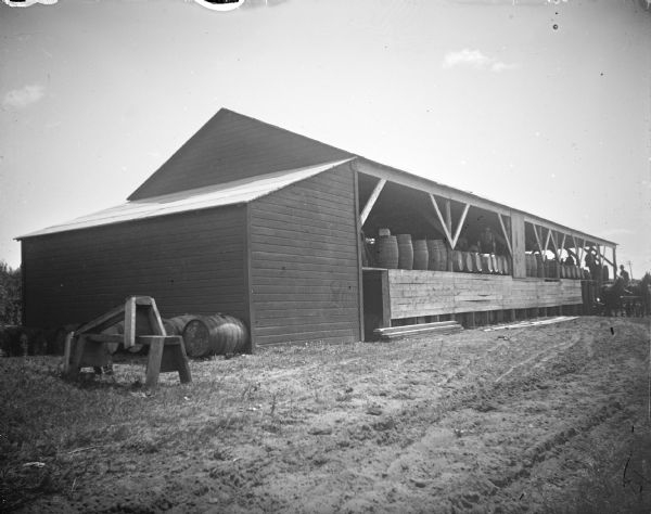 Outdoor view across yard towards several men inside a long building  with open sides showing a long line of barrels. In the background is a wagon next to side of the building. The building is identified as the Onalaska Pickle Factory one block north of the northeast corner of East Main (Highway 54) and Winnebago Street, on the edge of the tracks.