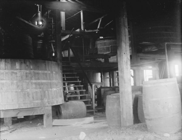 Indoor view of a wooden building with two levels. There is a set of wooden stairs leading up to the next level. Several barrels are near the base of the stairs. The building is identified as the Onalaska Pickle Factory one block north of the northeast corner of East Main (Highway 54) and Winnebago Street, on the edge of the tracks.