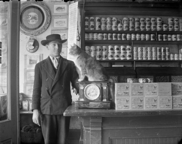 Interior portrait of a young European American man posing standing in a general store. He is wearing a dark-colored suit coat and hat, and is standing at the end of a counter, near a cat sitting on a clock on the counter. There are shelves of canned goods on the right, and the store entrance, with glass doors, is on the left. Arm & Hammer Soda trading cards of fish are hanging on the wall in the background.