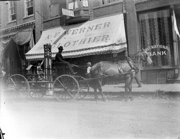 Outdoor view from street towards a European American man posing sitting on a wagon drawn by a single horse at the curb in front of A.F. Werner, The Clothier, storefront, and on the right the First National Bank. They are both decorated with patriotic banners with stars and bars.