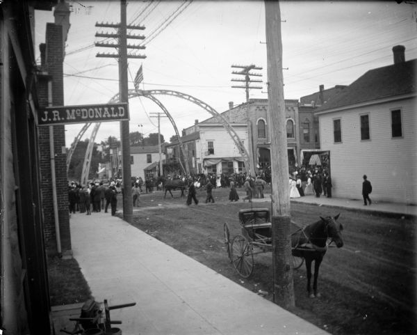 Slightly elevated view of a street scene looking north. There is a parade at the intersection of Main and First Streets.
