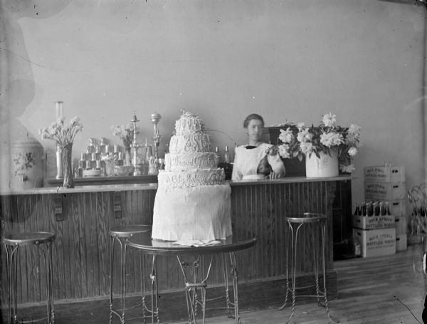 Indoor view of a European American woman posing standing behind a high shop counter. In front of the counter is a large decorated cake that reads: "Grand Opening, City Bakery." The shop is located on the north side of Main Street between First and Second Streets.