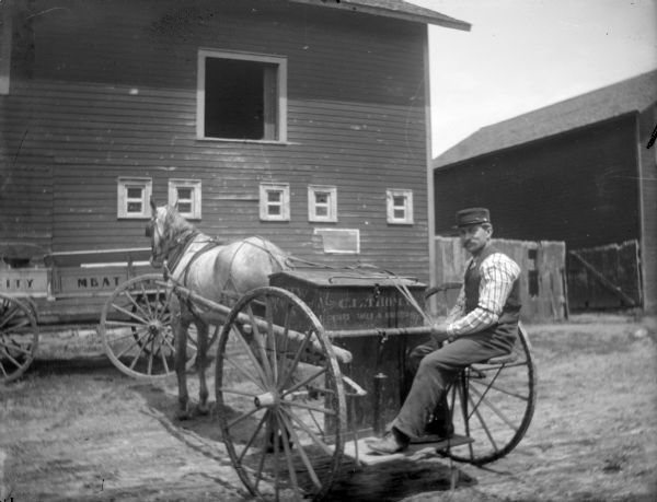 Outdoor view of a European American man posing sitting on the back of a horse-drawn delivery wagon marked "C. L. Thomas, Orders taken and delivered," pulled by a single horse. There is another wagon in the background parked near a wooden building.