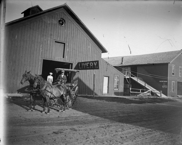 Outdoor view across road towards a man posing sitting in a wagon pulled by a pair of horses in front. In the background a woman is posing standing in the doorway of the Sawyer Livery and a paint shop. Probably on the east side of First Street between Main and Fillmore Streets.