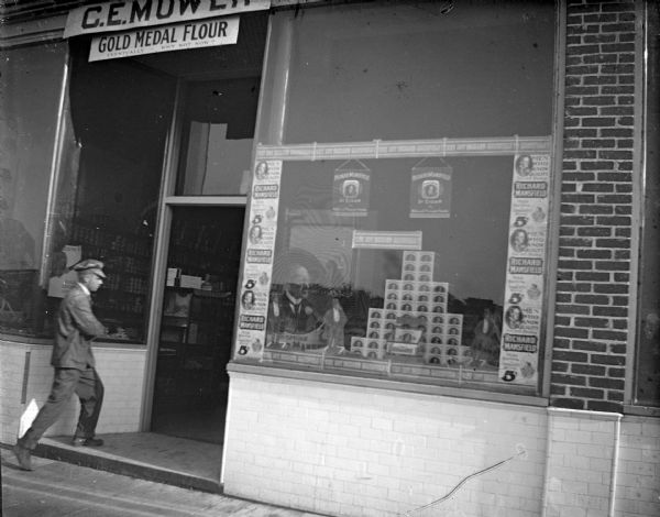Outdoor view of a European American boy wearing a cap who is entering the shop of C.E. Mower. Probably located on the north side of Main Street. There is a display of Richard Mansfield 5 cent cigars in the window.