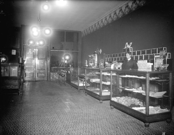 Indoor portrait of a European American man posing standing behind glass display cases in a shop with electric light bulbs in fixtures handing from the ceiling. Identified as the City Bakery, located in the third building east from the Second Street corner on the north side of Main Street.