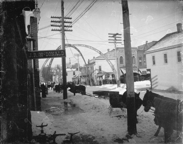 Outdoor view from snowy sidewalk of a horses and sleighs on a snow-covered First Street, looking towards its arched intersection with Main Street. Some of the horses are wearing blankets. There is a sign for J.R. McDonald on a building on the left.