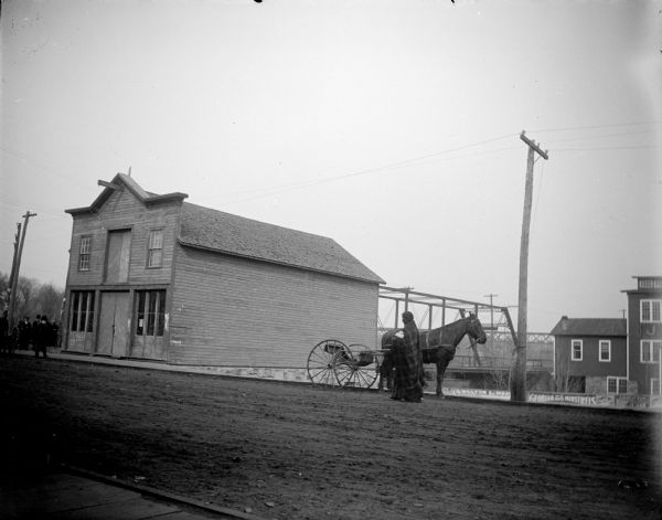 View across unpaved road towards a Native American woman, wrapped in a plaid shawl, walking in front of a horse hitched to a wagon. On the left across the street is a building with a large group of men standing on the sidewalk in front. The location is  identified as the southeast corner of Water and Harrison Streets.