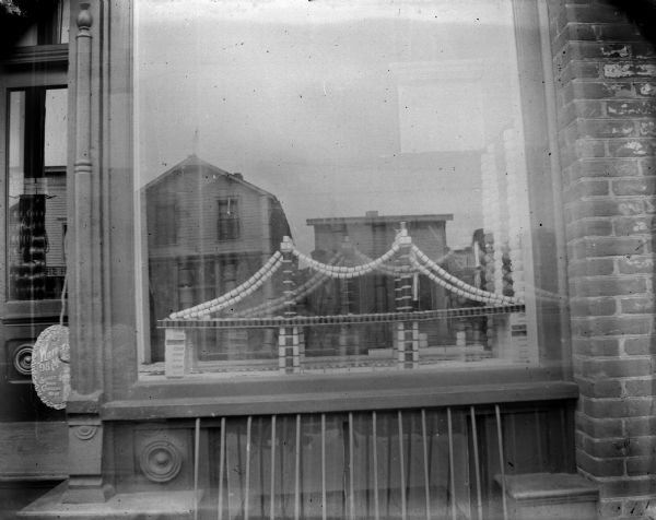 Outdoor view from sidewalk of a storefront window, possibly of the Werner Store, displaying products in the shape of a bridge. There is a sign on the left that reads: "Warner's 98 Model, Select Your Corsets Here." There are reflections of other buildings in the window, as well as of the photographer.