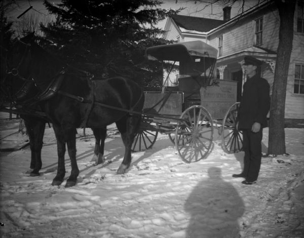 Outdoor portrait of a man posing standing next to a wagon pulled by two horses on snow-covered ground. The sign on the wagon reads: "Rawleigh's, Spices, Extracts. Every Article." The shadow of the photographer is in the foreground.