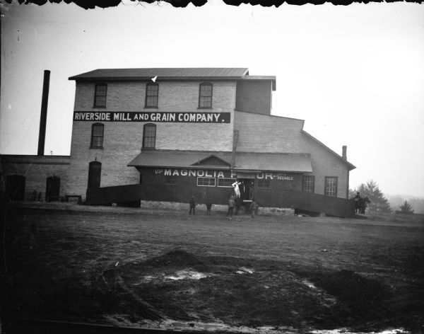 View across road of men and children posing standing in front of a large three-story brick building, with signs that read: "Riverside Mill and Grain Company" and "Use Magnolia Flous, Allways [sic.] Reliable." Identified as the Riverside Mill and Grain Company, located on highway 12, across the Black River and just south of the bridge facing east.