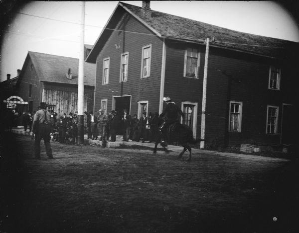 View across road towards a group of men standing on a sidewalk watching a man riding a horse in front of Building No. 88 on the northeast corner of South First and Fillmore Streets. Gruber reportedly had a paint store on the upstairs of Building No. 88.