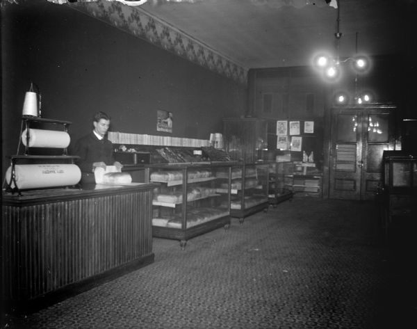 Interior view of a bakery, with a man posing standing behind a counter wrapping two loaves of bread. The bread is displayed in glass cases. Identified as the City Bakery.