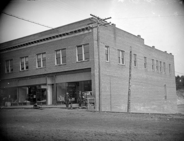 View across unpaved road towards a two-story building, with three men posing standing on the walkway. Identified as Moe Hardware, built in 1912 on the northwest corner of Main and First Streets.