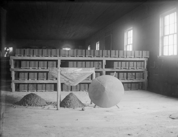 Indoor view of a cement factory showing shelves with molded cement blocks and other materials. Identified as the Mattson Cement Block Factory in Brockway.