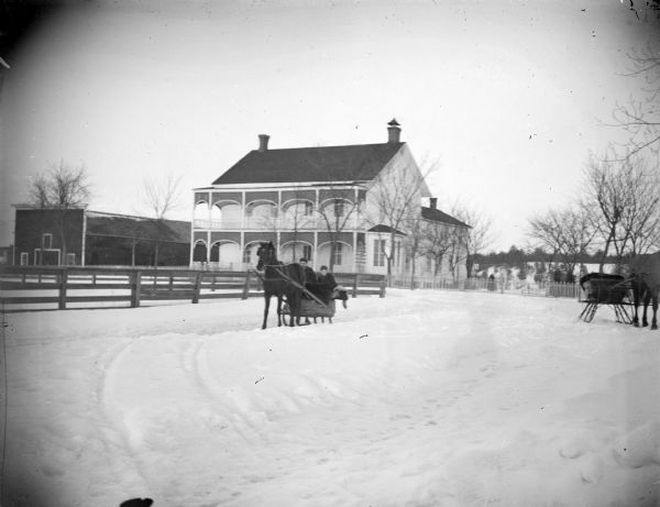 Outdoor view across snowy road towards a two-story wooden building surrounded by snow. In the middle foreground are two horse-drawn sleighs, the one in the center has two people. Behind them is a building identified as the United States Hotel, 326 South Second Street, the home/hotel owned by H.A. Bright.