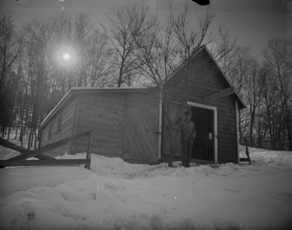 View looking up snow-covered hill towards a man posing standing next to a tree on a snow-covered ground in front of a wooden structure with a sign that reads "Blacksmith Shop."