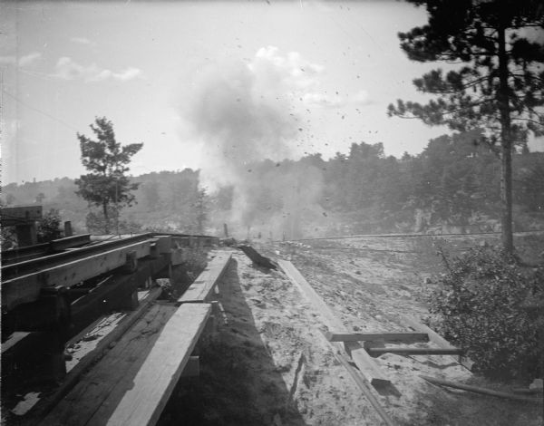 View looking down along wooden planks and machinery towards smoke in the distance. Identified as the rock crusher, located on the east side upriver from town, across from Spaulding's Rock and the cemetery.