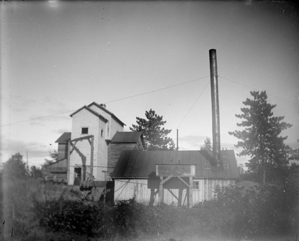 View across overgrown field towards a wooden structure with a tall smokestack. Identified as probably the rock crusher, located on the east side upriver from town, across from Spaulding's Rock and the cemetery.