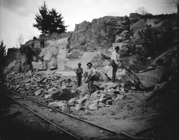 View across railroad tracks towards a group of men posing standing and sitting near a rock face and a track. Identified as probably the quarry connected to the rock crusher, located on the east side upriver from town, across from Spaulding's Rock and the cemetery.