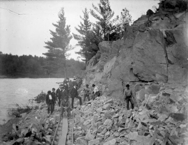 View down railroad tracks towards a group of men posing standing near and on a rock face and a track, with a river on the left. Identified as probably the quarry connected to the rock crusher, located on the east side upriver from town, across from Spaulding's Rock and the cemetery.