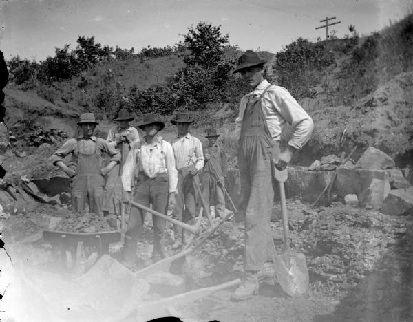 Group portrait of men posing standing near and on a rock face with shovels and pickaxes. Behind them are shrubs and trees on a hill, with a power line at the top. Identified as probably the quarry connected to the rock crusher, located on the east side upriver from town, across from Spaulding's Rock and the cemetery.