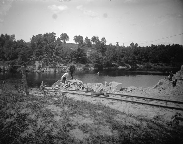 View across railroad tracks towards a group of men standing near and on a rock face an a cliff above a river. Identified as probably the quarry connected to the rock crusher, located on the east side upriver from town, across from Spaulding's Rock and the cemetery.