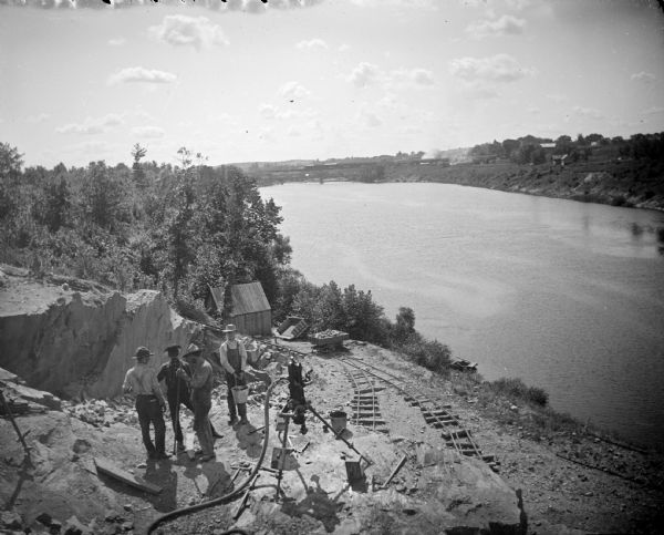 Elevated view of a group of men posing standing near and on a rock face with two sets of small railroad tracks behind them. Identified as probably the quarry connected to the rock crusher, located on the east side upriver from town, across from Spaulding's Rock and the cemetery. A railroad train is passing in the distance in the background on the opposite shoreline of the river.