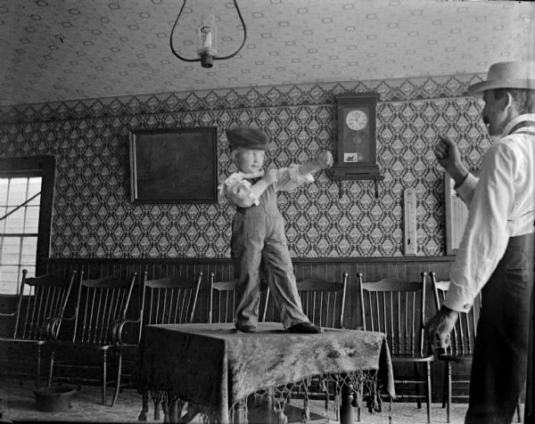 Indoor portrait of a European American child posing standing on a table in a pugilist pose, with a European American man posing standing on the right with a raised fist. They are inside the Merchant's Hotel.
