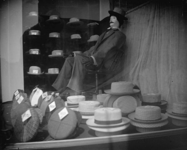 View of a window display of hats. Behind the hats is a mannequin sitting in a chair wearing a suit, hat, and a moustache. Behind the mannequin are shelves displaying more hats.