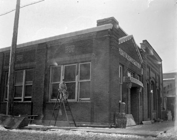 View from street towards a man posing sitting high up on a ladder set up on the sidewalk in front of the Jackson County Bank on the southwest corner of Main and First Streets.