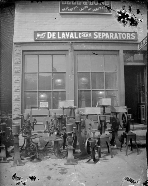 Outdoor view of a storefront with cream separators in front of a business. Identified as the Dell and Lund Real Estate Agency, which also sold farm equipment, including De Laval Cream Separators, located on south First Street, between the Van Schaick building and the Jackson County Bank.
