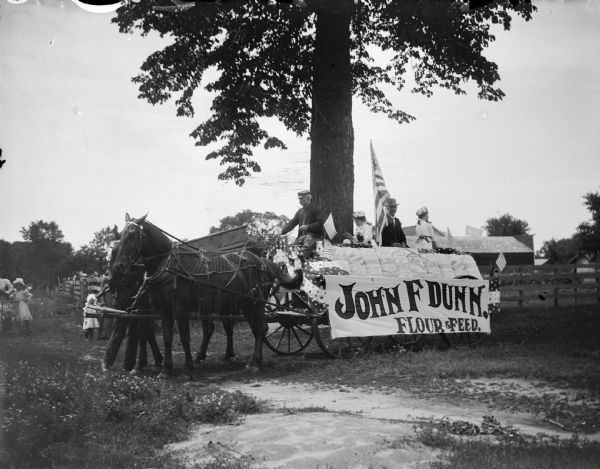 Outdoor view of a wagon with two men and two girls which is pulled by a team of two horse wearing fly nets, and decorated with a banner that reads "John F. Dunn, Flour and Feed."