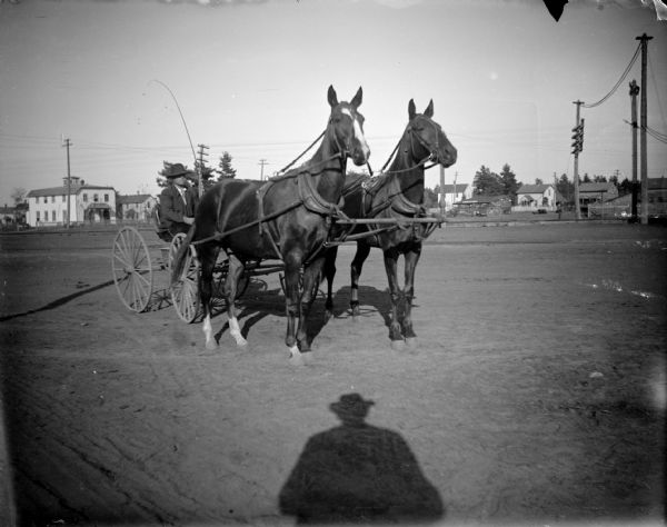 Outdoor portrait of a man posing sitting in a cart pulled by a team of two horses. Location identified as near the railroad depot looking northeast, with the Waldum House on the left side. The shadow of the photographer is in the foreground.
