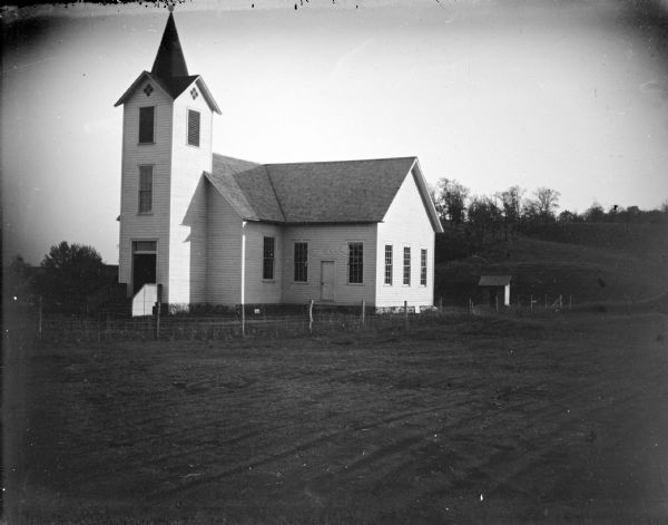 Outdoor view of a church surrounded by a wire fence near a hill. Identified as the Lutheran church near Squaw Creek.