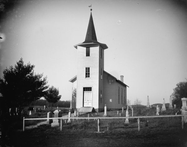 Outdoor view of a church surrounded by several grave markers. Identified as the Little Norway Lutheran Church.