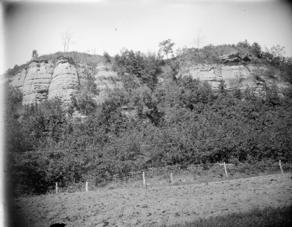 View across field and a fence towards a rock formation in a landscape surrounded by trees. Location identified as Castle Mound.