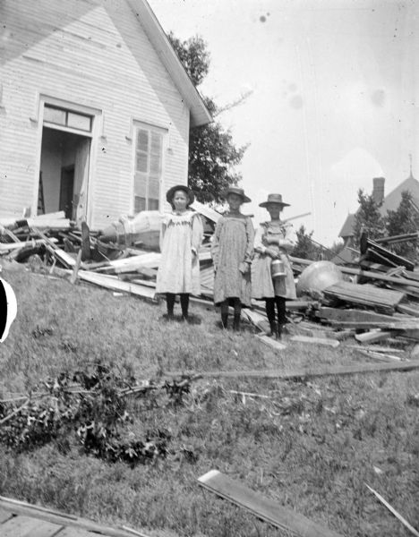 Outdoor group portrait of three girls posing standing in front of a church among wooden debris. Identified as the old Catholic Church (formerly a Universalist Reformed) located on the Main Street hill, whose steeple reportedly blew off in 1900. There's a bell in the debris behind the girls.