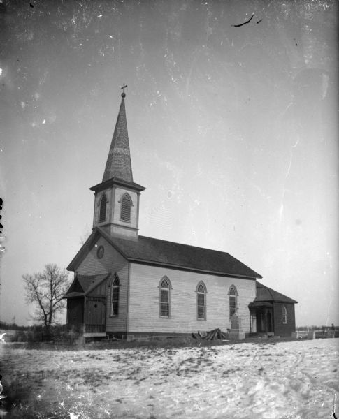 Exterior portrait of a church with a steeple and a few grave markers surrounded by a snow-covered ground. Identified as the St. Malachi Church in Trout Run.