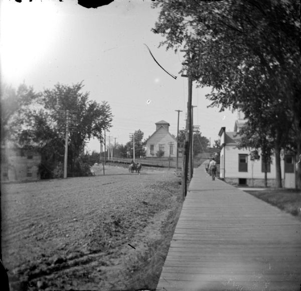 View down wooden sidewalk and unpaved street. There is a wooden church building on the hill in the distance. Identified as Main Street up the hill toward the old Catholic Church, which was reportedly torn down in 1941.