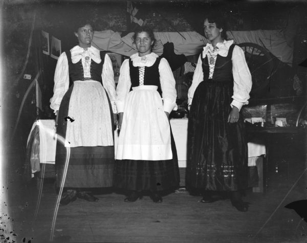 Indoor portrait of three European American women posing standing and wearing what is probably Norwegian costumes.